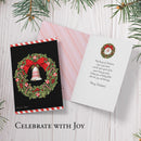 Christmas card with wreath design, envelope, and holiday message on a wooden surface with pine branches.